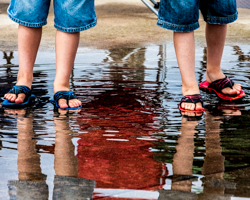 Twins must often negotiate their identity by contrasting their physical likeness with personal uniqueness. On this rainy summer day two brothers enjoy the reflective ripples and exuberant splashes of a bayside puddle. In the telltale details of their similar outfits individuality asserts itself in the stitches of their denim and the color variations of their flip-flops. Together they have choreographed in their water play a fraternal dance of youth.